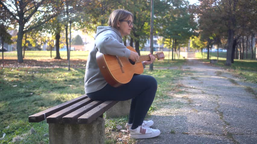 Beautiful woman sitting on bench playing acoustic guitar and singing alone outside in park. Happy Cheerful female enjoy sing some new sound on classical guitar Learning, practicing as hobby