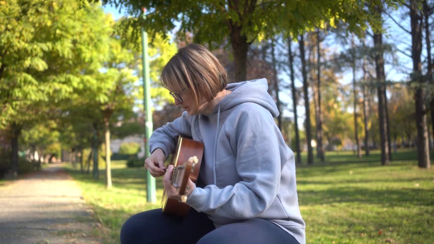 Beautiful woman sitting on bench playing acoustic guitar and singing alone outside in park. Happy Cheerful female enjoy sing some new sound on classical guitar Learning, practicing as hobby