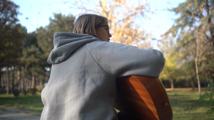Beautiful woman sitting on bench playing acoustic guitar and singing alone outside in park. Happy Cheerful female enjoy sing some new sound on classical guitar Learning, practicing as hobby