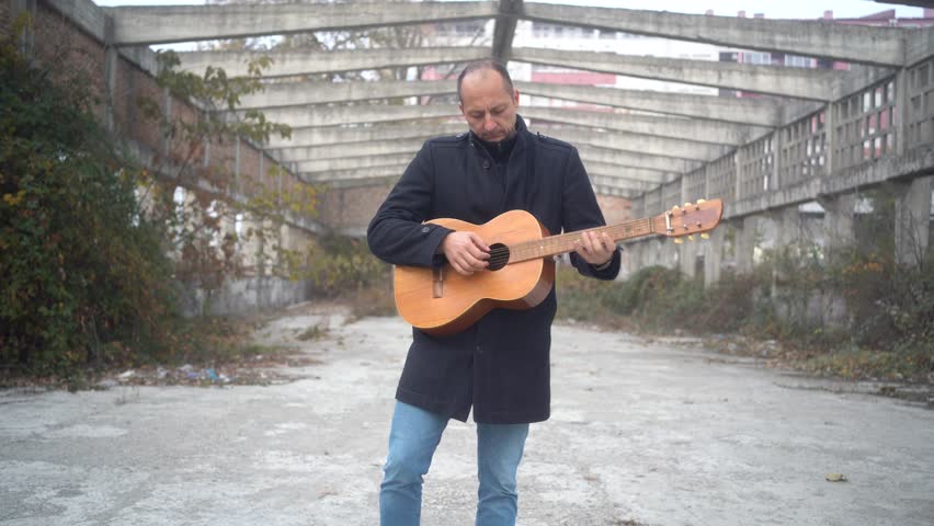 A man plays the guitar in an abandoned hall during autumn. The atmosphere is quiet, with no signs of life around him, adding to the feeling of isolation.
