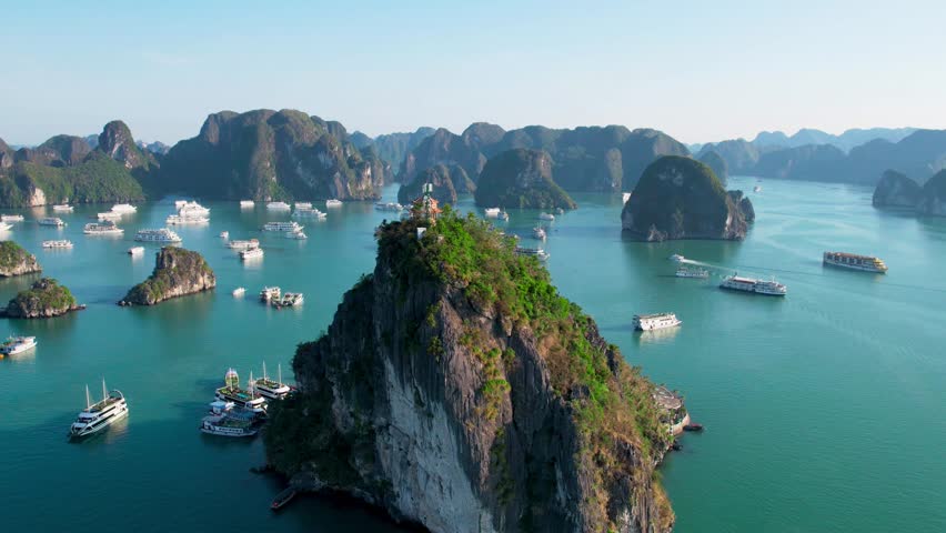 Aerial drone hyperlapse orbiting mountain with building in Ha long bay Cat Ba national park showing cruise ships and boats in emerald green waters of pacific with towering limestone pillar islands