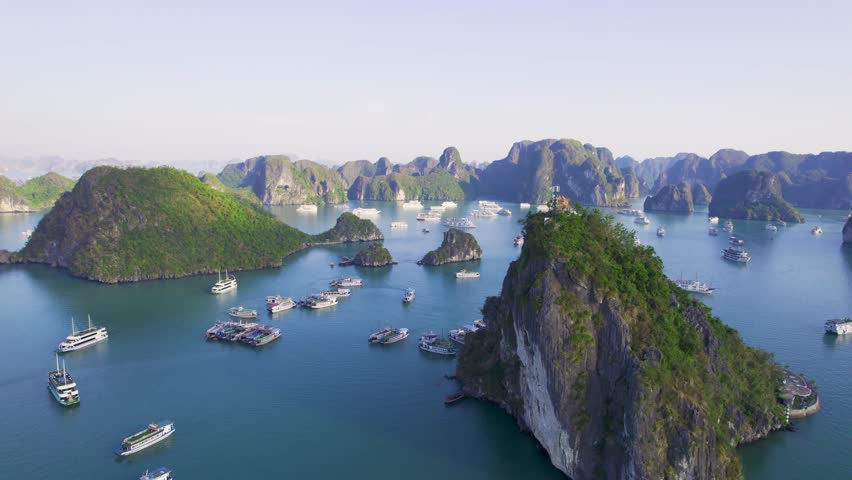 Aerial drone orbiting mountain with building in Ha long bay Cat Ba national park showing cruise ships and boats in emerald green waters of pacific with towering limestone pillar islands with trees