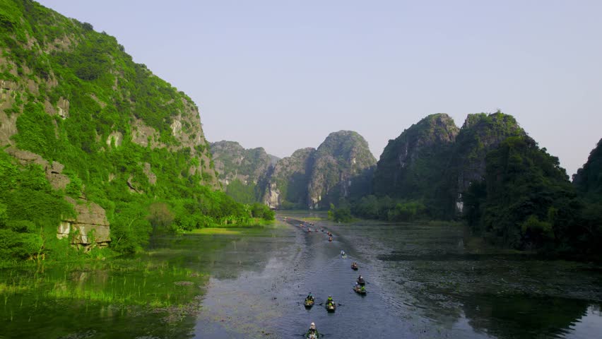 Aerial drone flying back over narrow river filled with traditional wooden rowboats surrounded by limestone cliffs mountains in Ninh Binh Vietnam