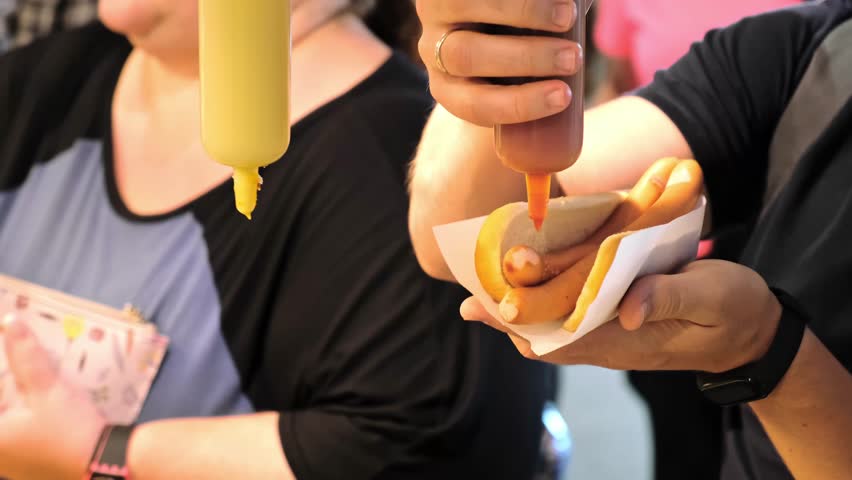 The man pouring ketchup on Hot Dog. Cooking of American Fast Food. Street food. Grilled Sausage on white bread bun. Catering self-service in airport. Fat person in queue in the background.