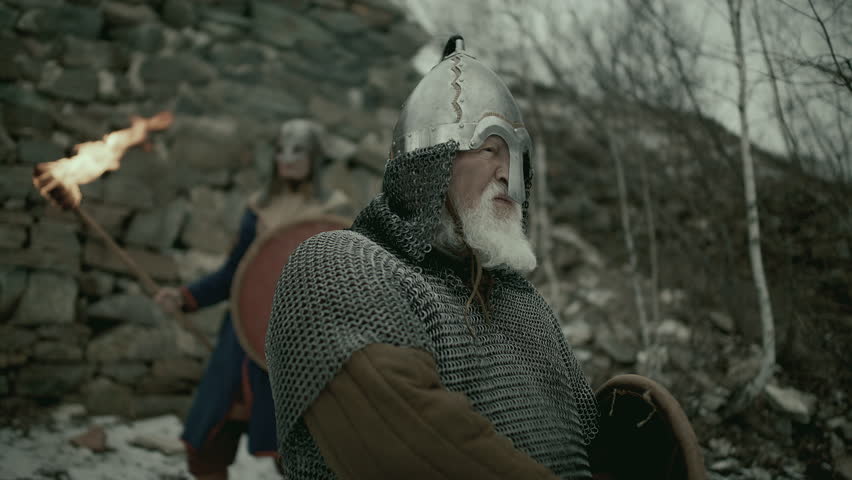 Senior bearded warrior wearing chainmail and metal helmet, holding sword and shield, shouting at the beginning of medieval battle by stone fortress
