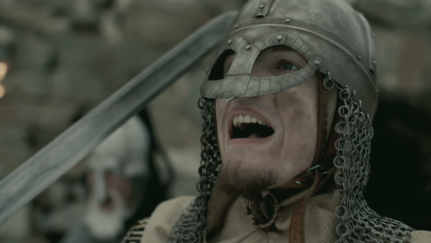 Medieval knight dressed in metal helmet and chainmail, holding sword and shouting at enemy on battlefield. Close-up view