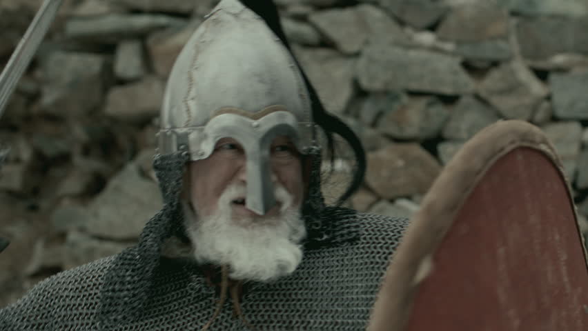 Grizzled warrior dressed in medieval chainmail and helmet, holding shield and striking with sword, fighting with enemy against stone wall of fortress