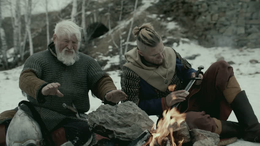 Elderly medieval warrior with white hair and beard sitting by fireplace, warming his hands above flames and talking to younger friend cleaning his sword during rest after battle in snowy landscape
