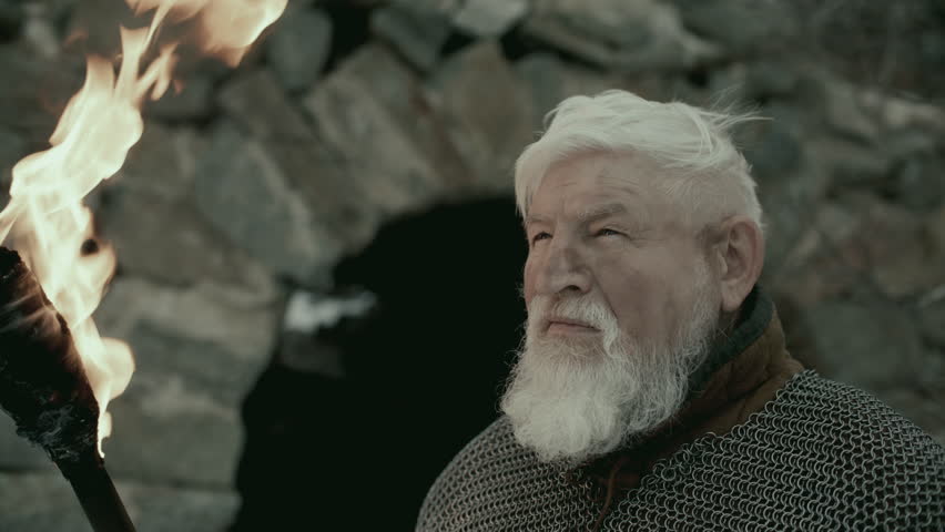 Elderly medieval guard with white beard wearing chainmail armor, standing outside ancient stone fortress on winter day, holding torch and gazing into distance