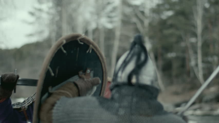 Medieval warrior in helmet and chainmail armour, holding shield and fighting opponent with sword outdoors in wintry rocky terrain