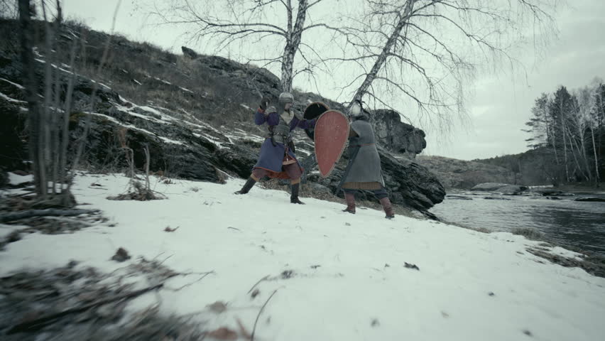 Two medieval warriors, dressed in chainmail, helmets and period clothing, fighting with swords on snow-covered ground near river and rocky hillside. Zoom shot