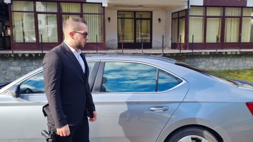 A professional bodyguard security officer in formal black attire surveys the surrounding opens the door for VIP. The scene suggests protection, private security, and executive transportation services.
