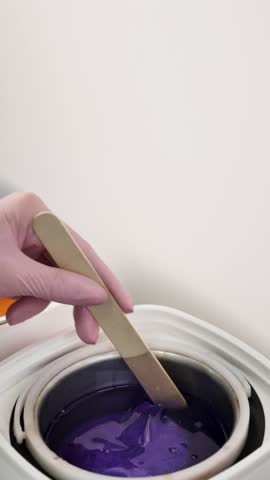 A depilatory master stirs purple granular wax into a wax melter with a wooden spatula, close-up, no face, vertical