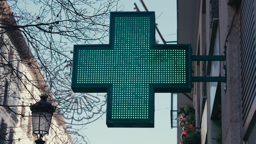 A vibrant green LED pharmacy cross sign stands out on a street corner in Madrid, Spain. The bright light indicates the presence of a nearby pharmacy, a common feature in European cities, symbolizing