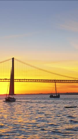 View of 25 de Abril Bridge famous tourist landmark of Lisbon connecting Lisboa and Almada on Setubal Peninsula over Tagus river with passing tourist yacht boat silhouette on sunset. Lisbon, Portugal