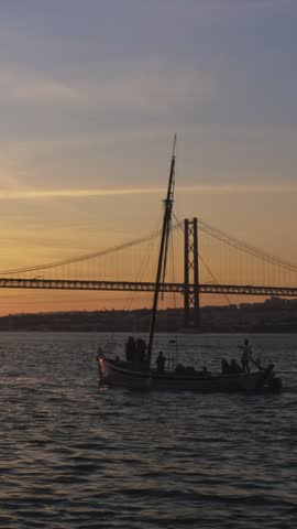 View of 25 de Abril Bridge famous tourist landmark of Lisbon connecting Lisboa and Almada on Setubal Peninsula over Tagus river with passing tourist yacht boat silhouette on sunset. Lisbon, Portugal