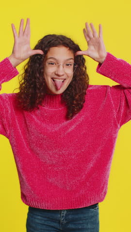 Caucasian teenager girl in blouse showing tongue making faces at camera, fooling around, joking, aping with silly face, teasing, bullying, abuse. Young teen child isolated on yellow background indoors