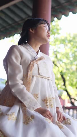 A Korean woman in her 20s wearing a Hanbok sits in a historical building in Seoul, South Korea. Vertical slow motion video.