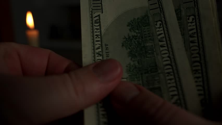 A man counts several dollar bills under the dim light of a candle, reflecting on high utility bills and the ongoing energy crisis, amid rising prices affecting many households.