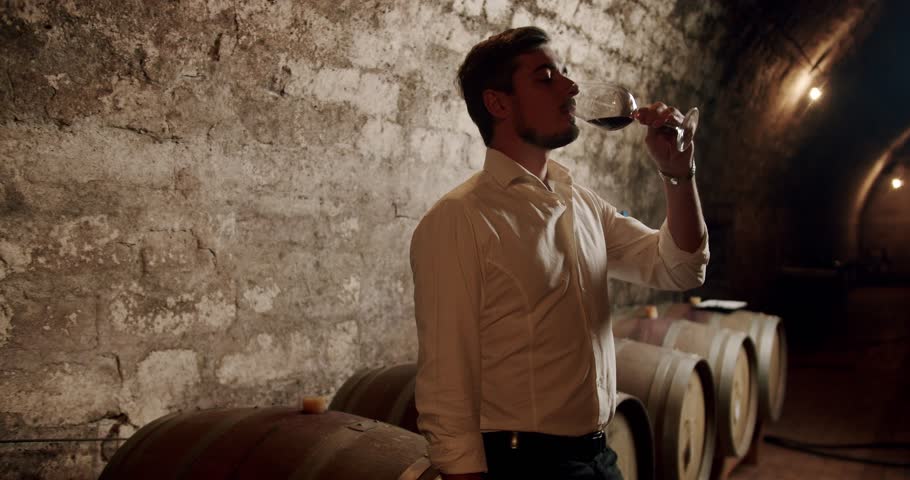 Happy successful male winemaker is tasting a flavor and checking fresh red wine quality poured in transparent glass in a wine cellar. Nicely dressed Sommelier checking and drinking a glass of red wine