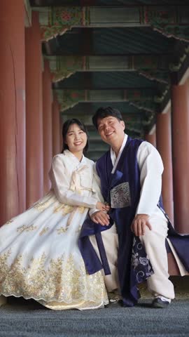 A Korean couple, a man in his 30s and a woman in her 20s, wearing Hanbok from Seoul, South Korea, sit side by side in a historical building. Vertical slow motion video.