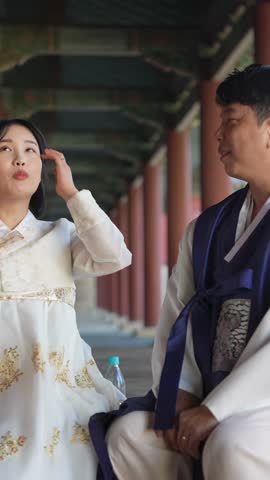 A Korean couple, a man in his 30s and a woman in her 20s, wearing Hanbok from Seoul, South Korea, sit side by side in a historical building. Vertical slow motion video.