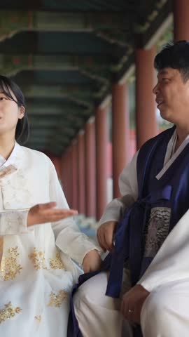 A Korean couple, a man in his 30s and a woman in her 20s, wearing Hanbok from Seoul, South Korea, sit side by side in a historical building. Vertical slow motion video.