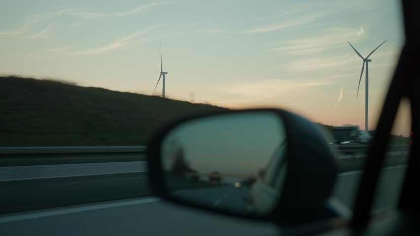 View of wind turbines at sunset through car window and side mirror with blurred highway in foreground - Powered by Shutterstock - Get 15% off with code: PIKWIZARD15