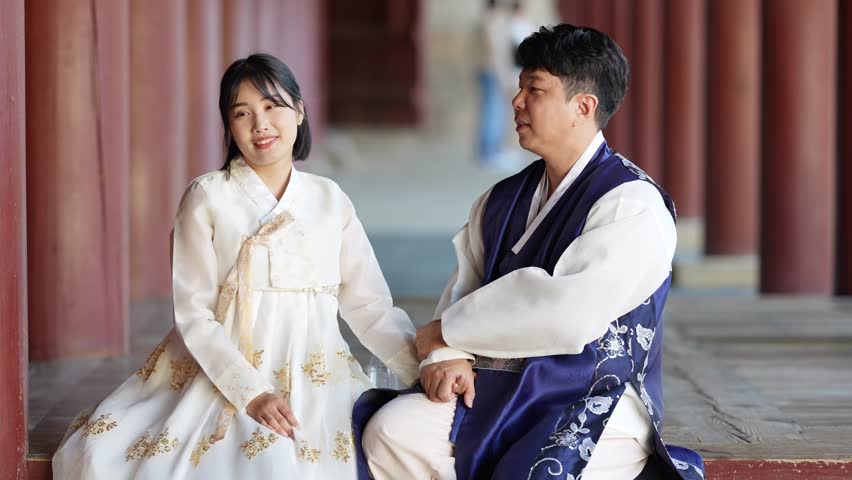 A Korean couple, a man in his 30s and a woman in her 20s, wearing hanbok from Seoul, South Korea, are sitting in a historical building, drinking water and using their smartphones.