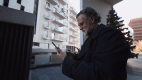 mature Caucasian businessman, around 50 years old, smiles warmly while looking at his smartphone, receiving notification. Dressed in a black coat, he stands outdoors and using mobile banking app - Powered by Shutterstock - Get 15% off with code: PIKWIZARD15