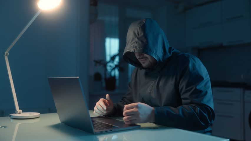 A frustrated hacker sits at a table in a dimly lit room with his laptop, expressing anger and stress after a failed attempt in his dark home environment.