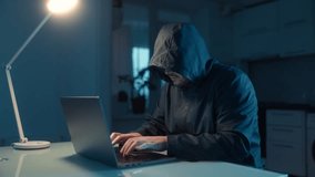 A hooded hacker intensely typing on a laptop in a dark room at home, expressing anger and frustration while sitting at a table, showing failure and stress. - Powered by Shutterstock - Get 15% off with code: PIKWIZARD15
