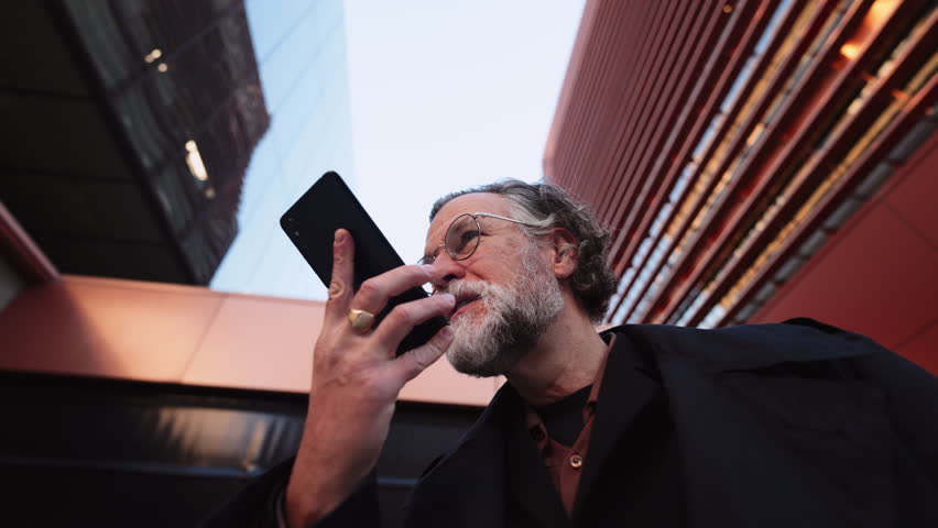 60 years old man records voice message on smartphone, surrounded by sleek modern architecture. low-angle view, integration of technology into everyday life communication.Sending audio to colleague