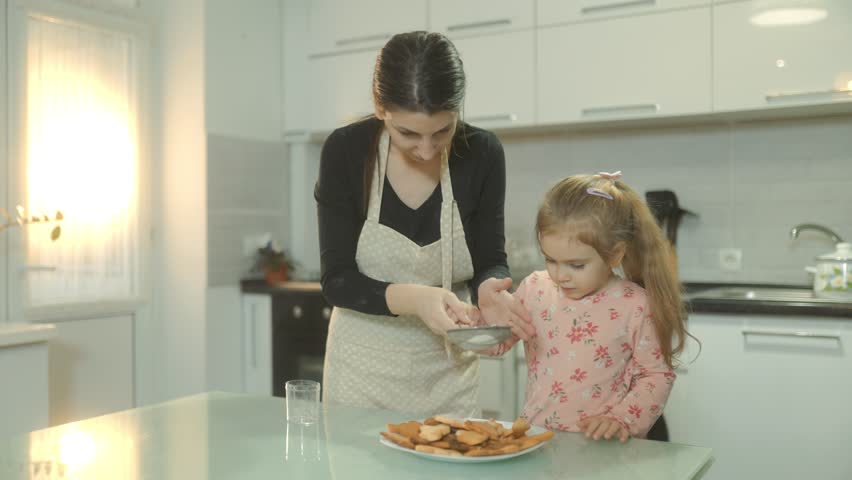 A mother and her young daughter bond in the kitchen as they sprinkle powdered sugar over freshly baked homemade biscuits. A warm and creative family activity at home.