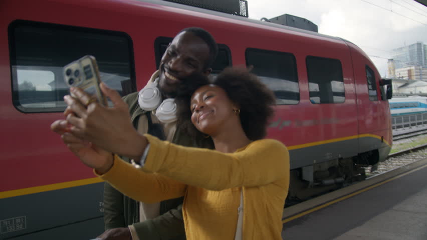 Joyful African American couple taking a selfie at a railway station before embarking on their train journey together, medium shot. Travel happiness concept.