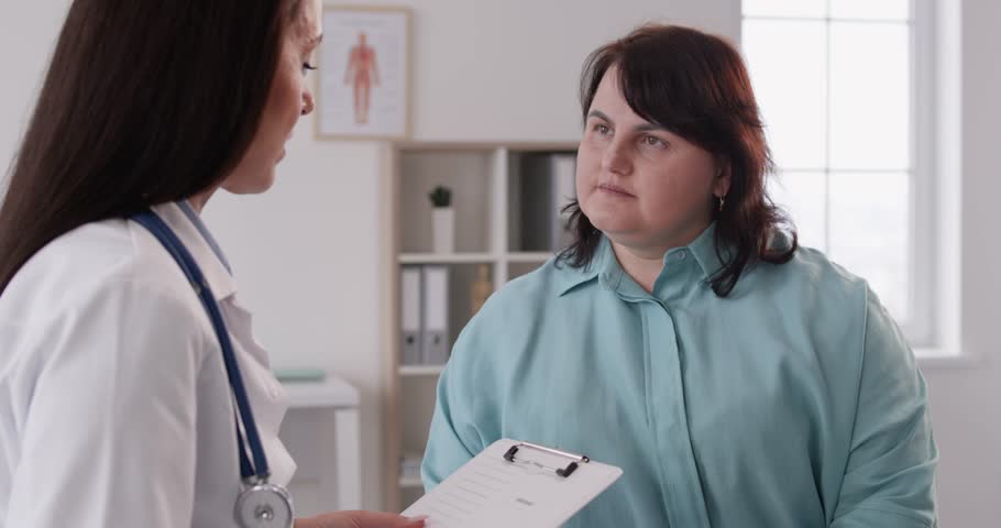 Portrait of fat overweight female patient in medical office listening to a friendly doctor holding report file with appointment and giving consultation a woman during medical exam in clinic. 4k video