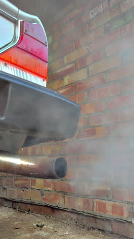 Vertical video - Closeup of smoke fumes pollution belching from the rear exhaust tail pipe of an old classic vintage car, standing in a brick garage.