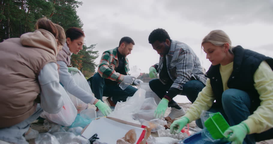 Group of people helping nature recover. Cleaning pollution in sand beach of big lake or river. Actively picking up waste material and sorting everything. Wearing gloves for hygiene.