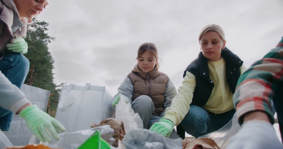 People collectively picking up pieces of plastic from ground and placing trash into big white bags. Charming little girl stopping while looking at camera and smiling. Cold weather.