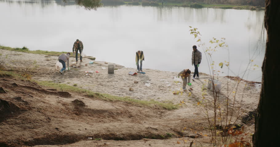 Camera view from above of group of people working together. Collectively cleaning sand beach of lake or river. Picking up pieces of plastic left or dropped by others. Assisting nature.