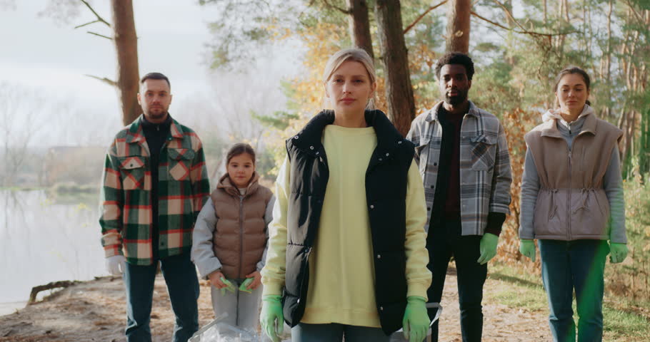 Volunteers standing together in forest. Sun shining bright. Looking at camera with focused expression. Wearing green gloves. Finishing picking up plastic waste. Chilly autumn weather.