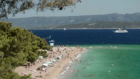 Crowds of tourists sunbathe, swim, and relax on Zlatni Rat Beach on the island of Brac (Brač), Croatia. A vacation on the Adriatic Sea coast during the tourist season. Waves crash against the shore. - Powered by Shutterstock - Get 15% off with code: PIKWIZARD15
