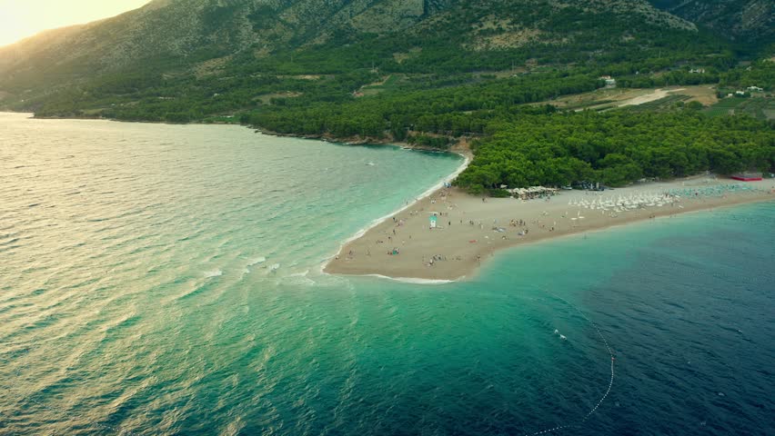 Zlatni Rat beach at sunset. People swim in the sea, swinging on the waves and watching the sunset, relaxing and sunbathing on the shore. Tourism and beach recreation on the Adriatic coast of Croatia.