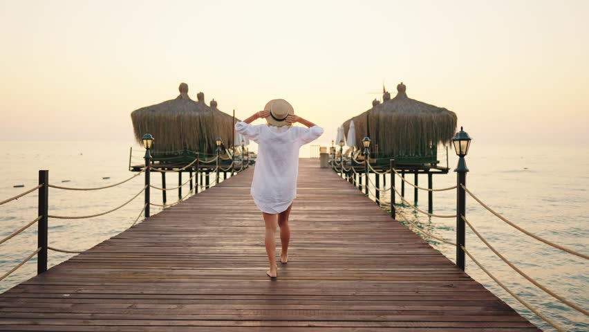 Slender blonde woman in white cotton shirt walking alone on wooden pier. Beautiful sunset or sunrise and female person in marina, back view, following shot, slow motion, carefree lady traveler