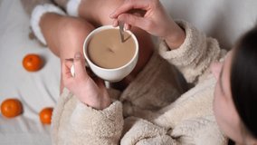 Woman in a bathrobe is sitting on a white bed, stirring a cup of coffee with a spoon. There are tangerines scattered on the bed around her - Powered by Shutterstock - Get 15% off with code: PIKWIZARD15