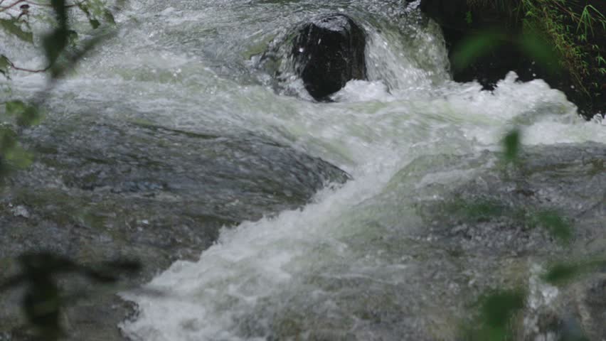 Cascades of natural torrent flowing between rugged rocky surface with green forest foliage in the foreground.
