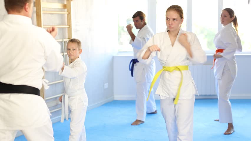 Teen girl with her family in kimonos and colored belts practicing karate with punches during group martial arts class in gym, accompanied by trainer