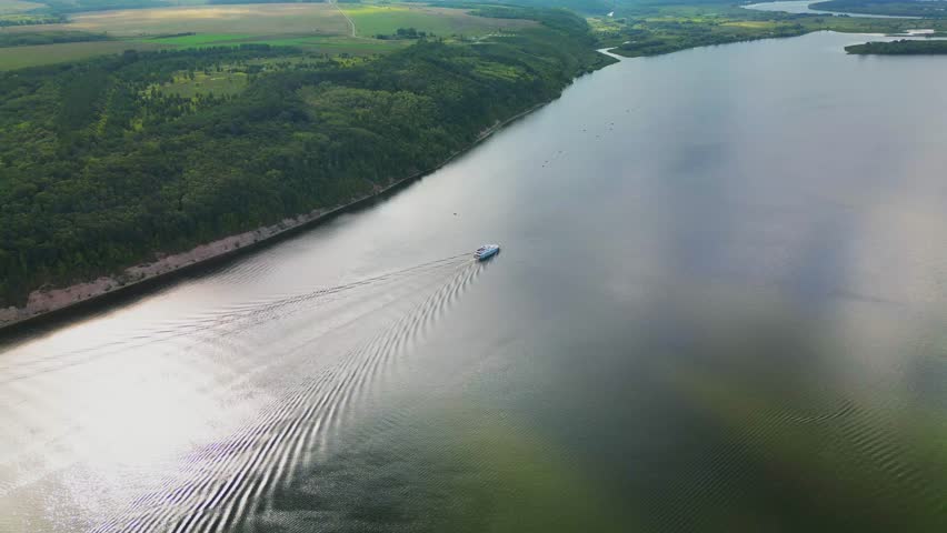 the ship goes through the water of the river forming waves. Top view of the wide river, forest and fields