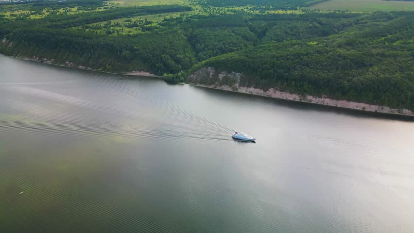 the ship goes through the water of the river forming waves. Top view of the wide river, forest and fields