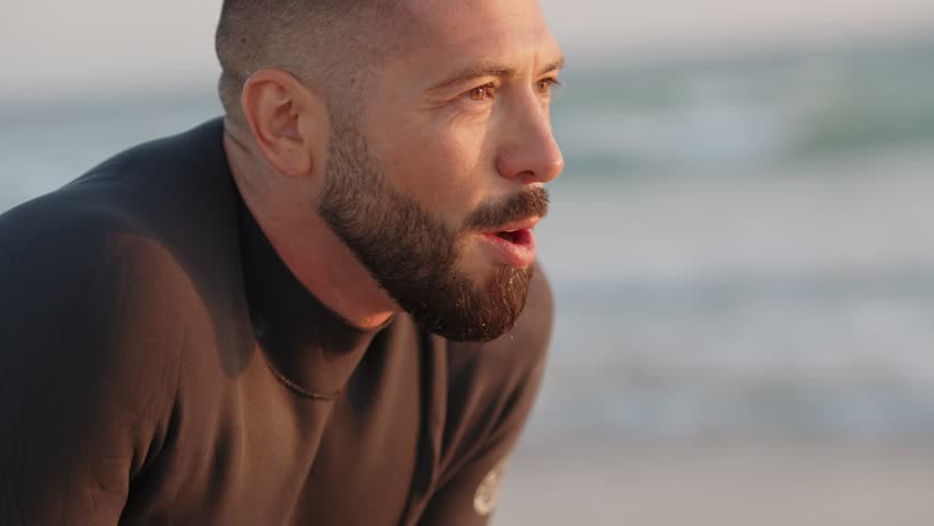A tired surfer catches his breath after a surf session on the beach at sunset. He
